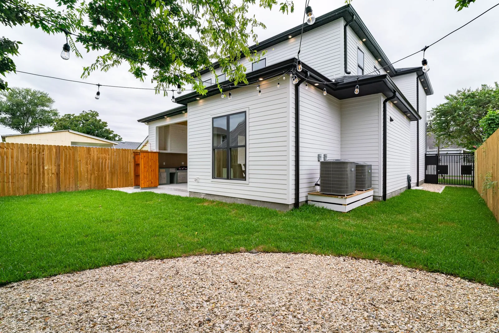 Two central air conditioner condensers on a raised wood pad beside a white-sided two-story house, green summer lawn, wood privacy fence and leafy trees in the backyard