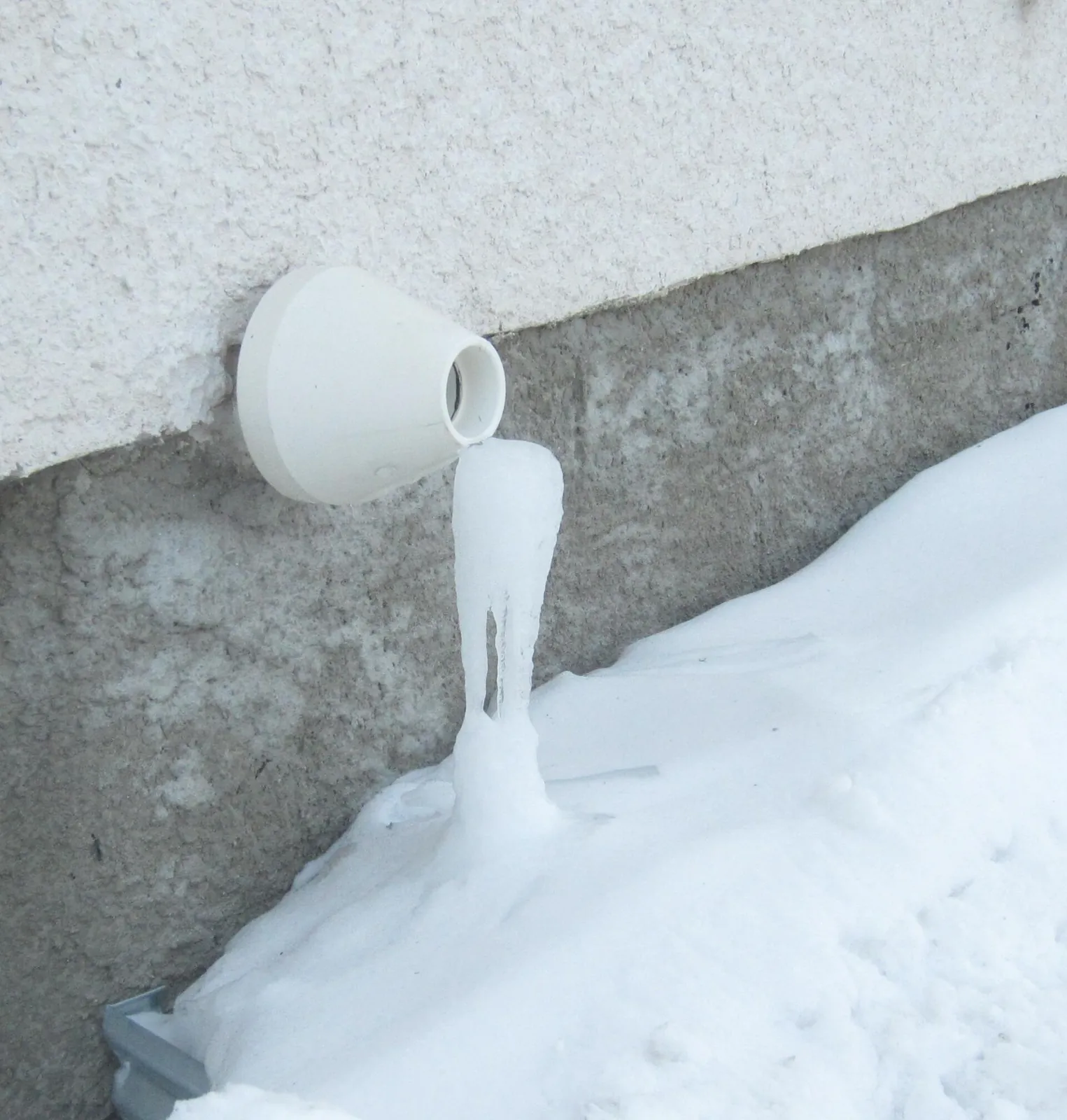 White PVC exhaust vent of a high-efficiency furnace on a stucco wall, with an icicle of frozen condensate running down into the snow