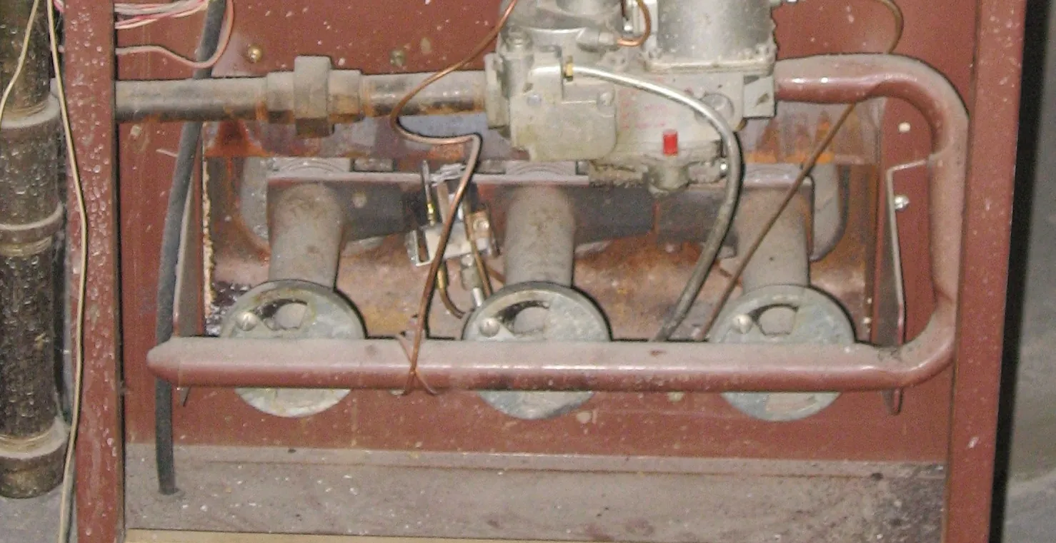 Older gas furnace with the burner door off, showing three burner tubes, the manifold pipe, pilot tubing and the gas valve