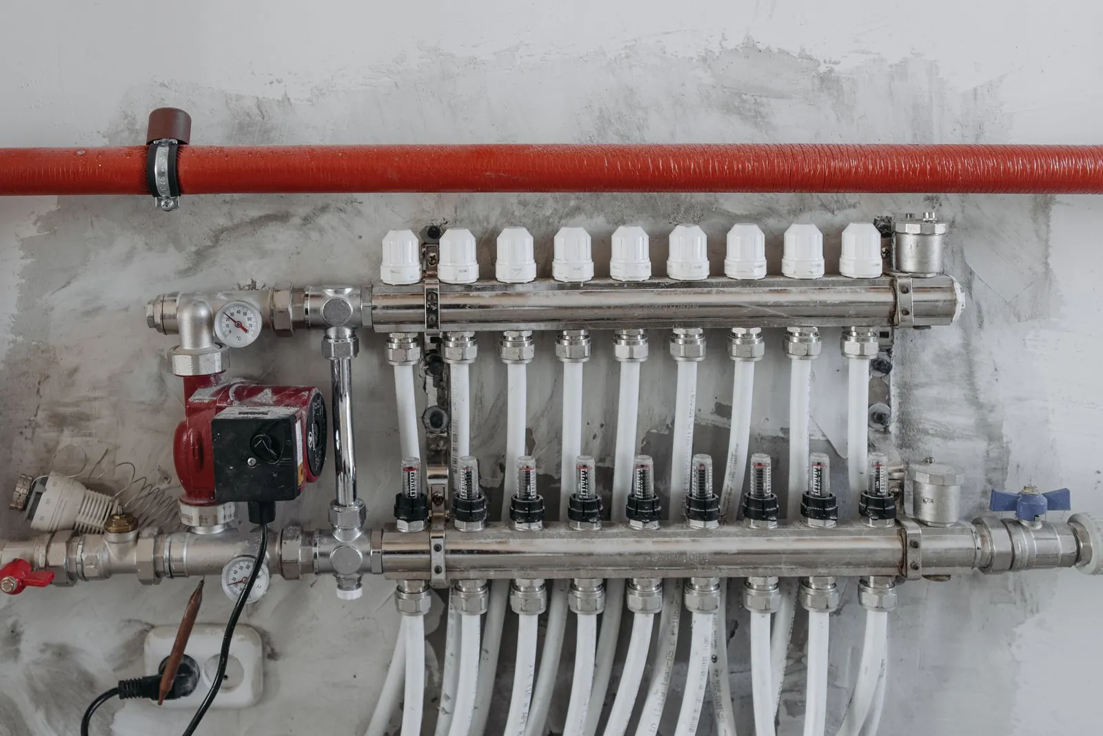 Hydronic radiant heating manifold with a red circulator pump, copper and steel supply piping and PEX loops on a basement wall