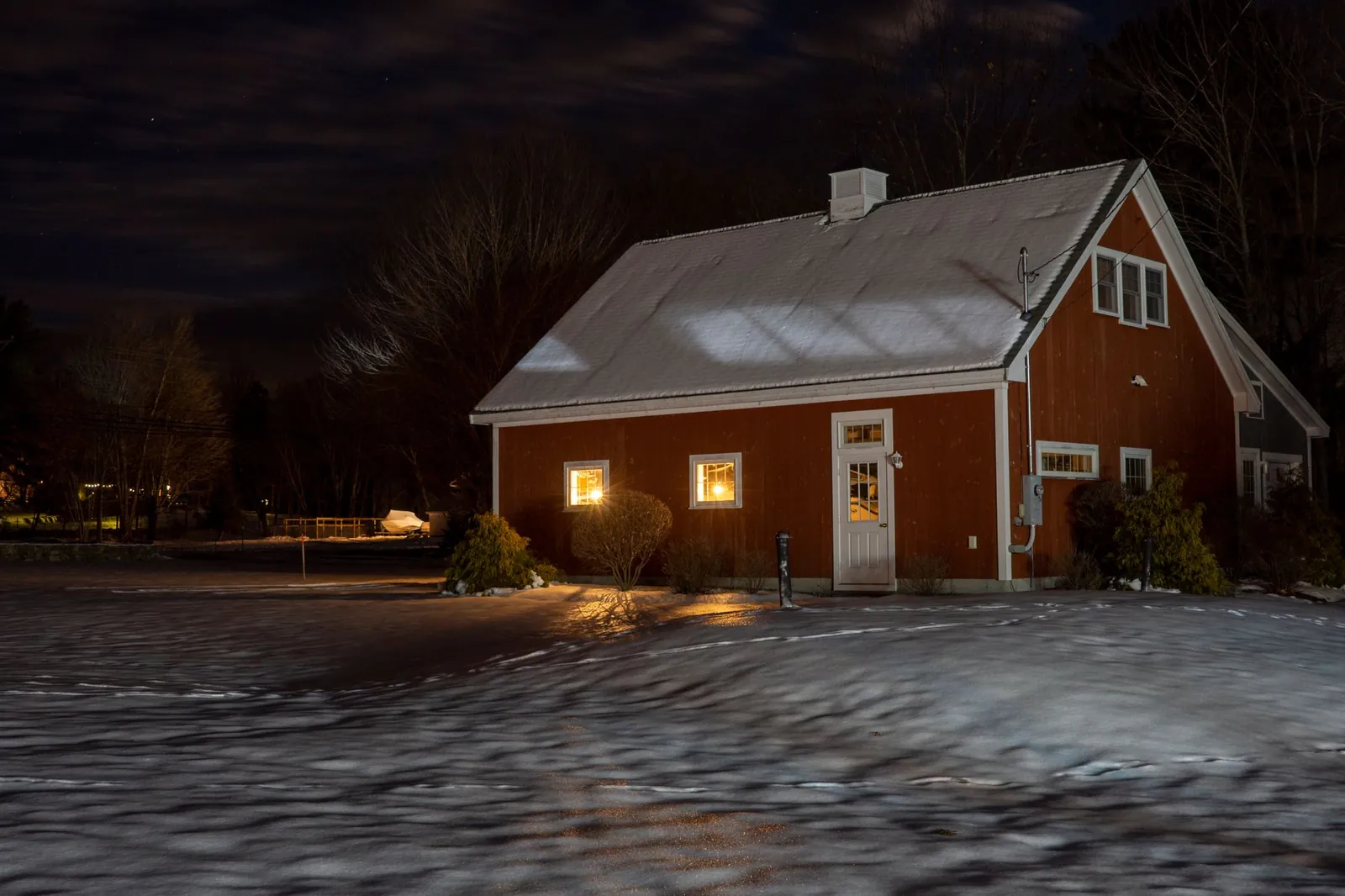 Red-sided house with a snow-covered roof and lit windows on a cold winter night, snow across the yard